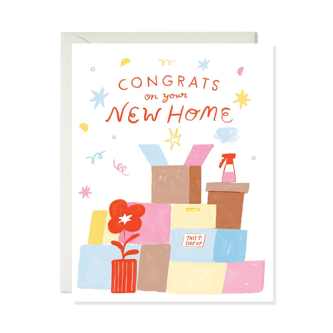 A white greeting card with a light gray envelope behind it, featuring a colorful hand-drawn illustration of stacked boxes, a red flower in a vase, and the text 'CONGRATS on your NEW HOME' in orange-red lettering.