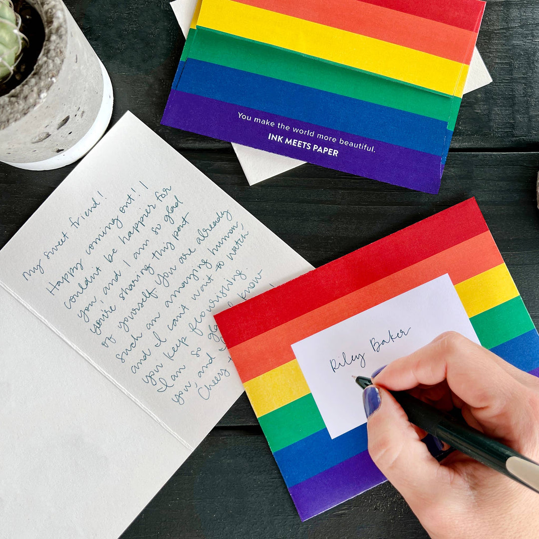 Open greeting card with hand-written text; hand holding pen addressing rainbow-striped envelope