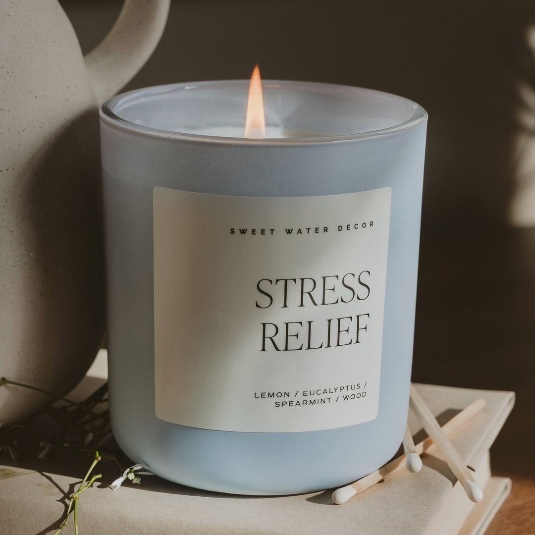 Lit candle in light blue matte glass container a square white 'Stress Relief' label.