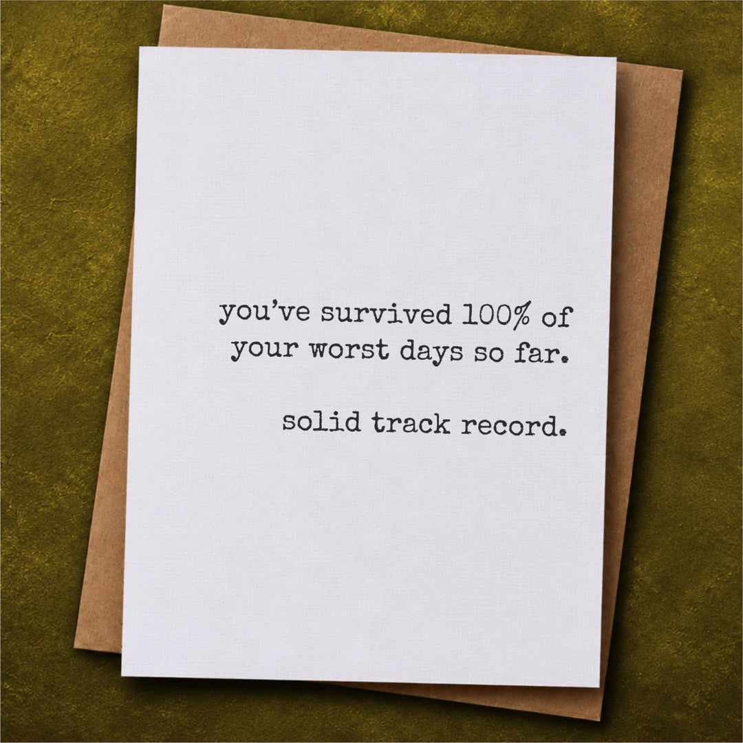A white greeting card with black typewriter-style text reading 'you've survived 100% of your worst days so far. solid track record.'