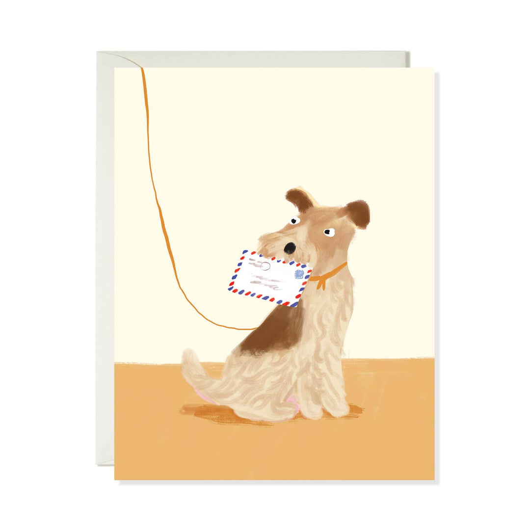 A greeting card featuring an illustration of a wirehaired dog sitting on a tan floor against a cream background, holding a postcard in its mouth with a leash attached. A white envelope is visible behind the card.