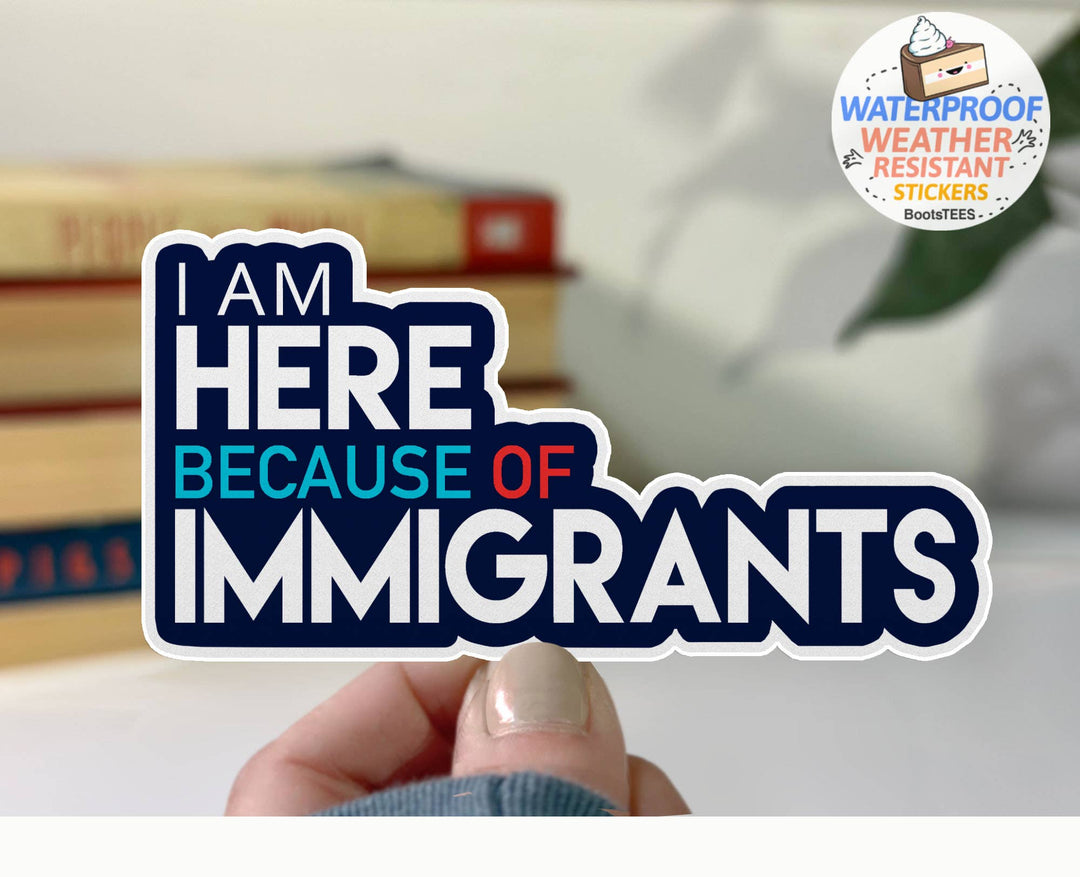 Die-cut sticker reading 'I AM HERE BECAUSE OF IMMIGRANTS' is held by a hand in front of a softly blurred indoor background with books and a plant. 