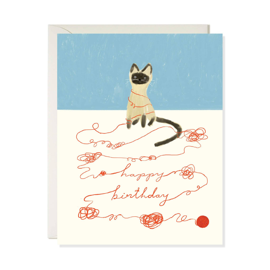 A greeting card with an illustrated Siamese cat on the pale blue upper half sitting beside red yarn-like script reading 'happy birthday' on an off-white lower half. An  off-white envelope is visible behind the card.