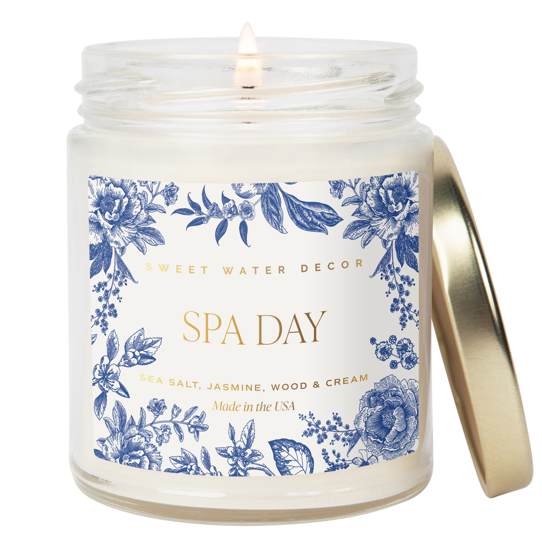 Lit candle in a white opaque jar with gold tone metal lid and gold text on a white label decorated with delft blue flowers.