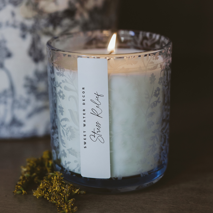 Lit candle in clear glass container with a floral print and a narrow white 'Stress Relief' label affixed vertically.