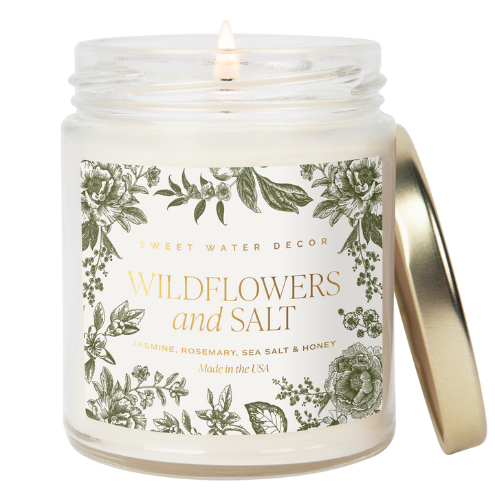 Lit candle is white opaque jar with  gold tone lid, and label with gold text and decorated with flowers,