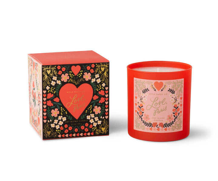 Candle in red glass container with label of a floral design with text 'Love, Paris,' in gold script, next to matching box.