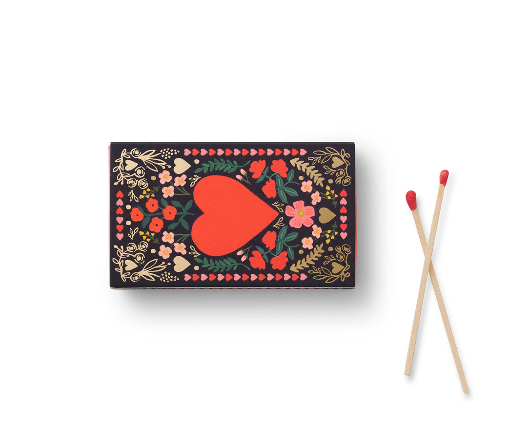 Two long matches with red tips, next to a closed matchbox with floral design and red heart in the center.