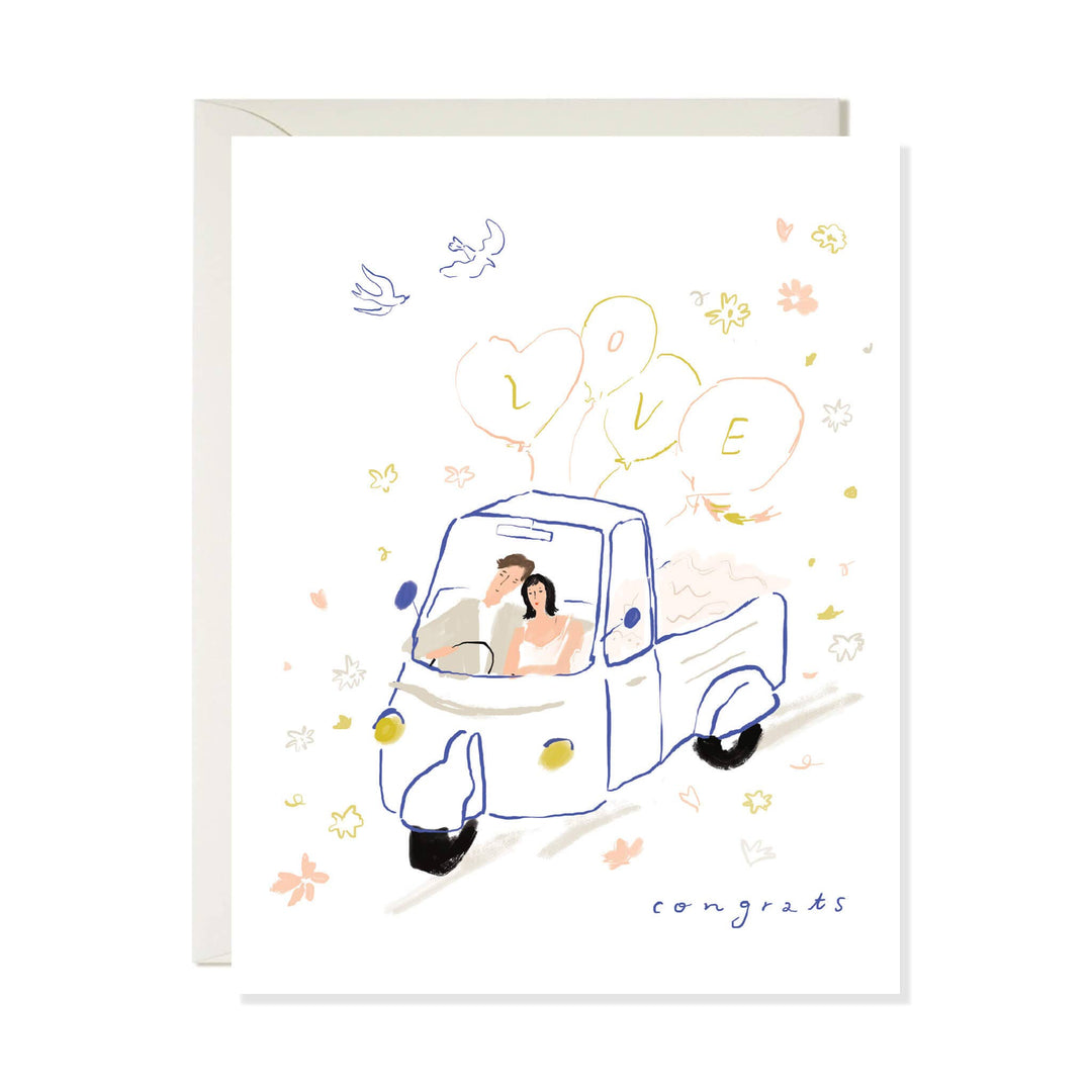 A white greeting card with a hand-drawn illustration of a couple in a small blue truck, surrounded by balloons spelling LOVE, flowers, birds, and the word 'congrats' at the bottom. A light beige envelope is placed behind the card.