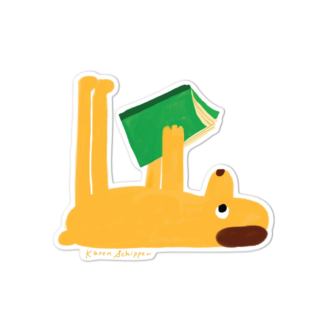 A die-cut sticker featuring a whimsical illustration of a yellow dog lying on its back and holding a green book overhead. The sticker has a white border and is signed 'Karen Schippen' in yellow script near the bottom left.