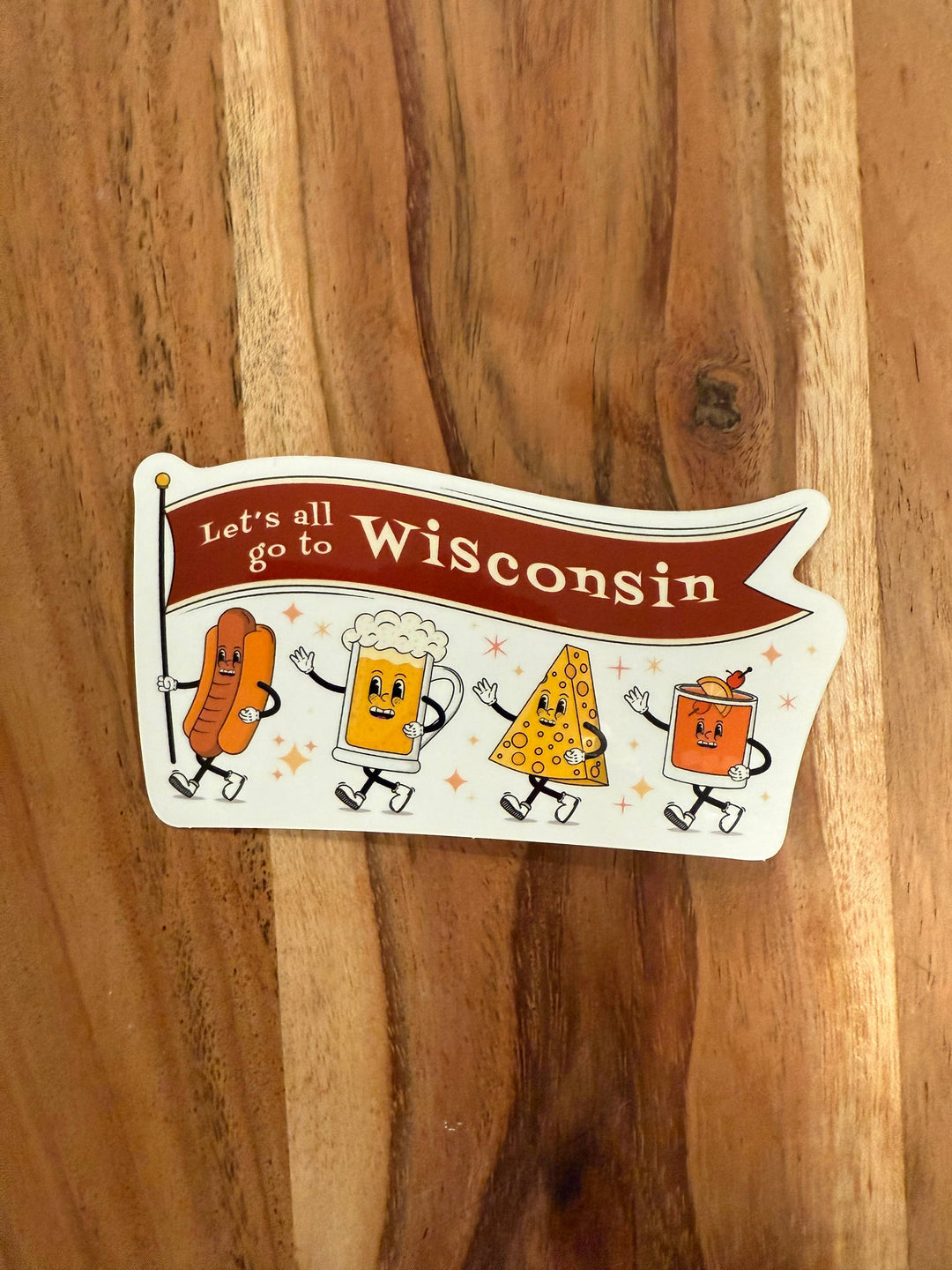 A die-cut sticker featuring a hot dog, mug of beer, cheese wedge, and old fashioned as cartoon characters holding a brown banner with the text 'Let's all go to Wisconsin' 