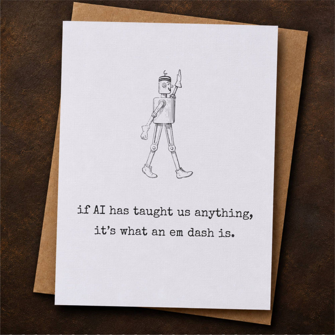 A white greeting card with a black line drawing of a walking robot and the text 'if AI has taught us anything, it's what an em dash is.' placed on top of a brown kraft envelope.