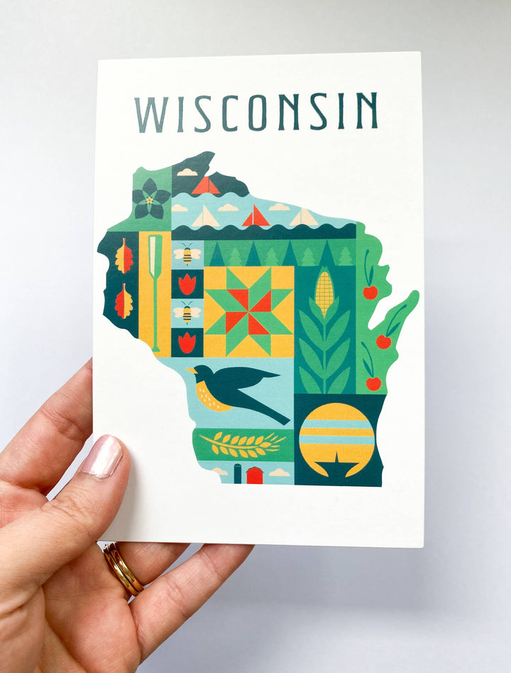 A hand holding an art print featuring a colorful map of Wisconsin filled with state-themed illustrations and the word 'WISCONSIN' at the top.