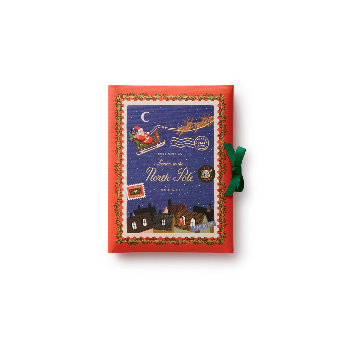 The folio for the stationery with a red cover that features Santa Claus in his sleigh against a starry night sky as he flies over a village, with a green ribbon closure.
