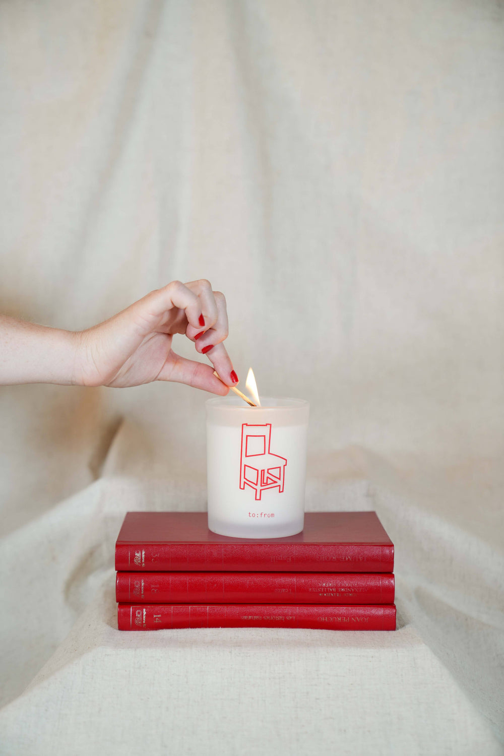 Matte white glass container printed with chair in red, sitting on three red books, being lit with a match.