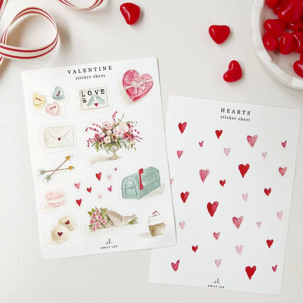 Two sticker sheets on a table with valentine heart candy. One sheet has watercolor dark and light red hearts in various sizes. The other sheet has a dozen valentine-inspired images.