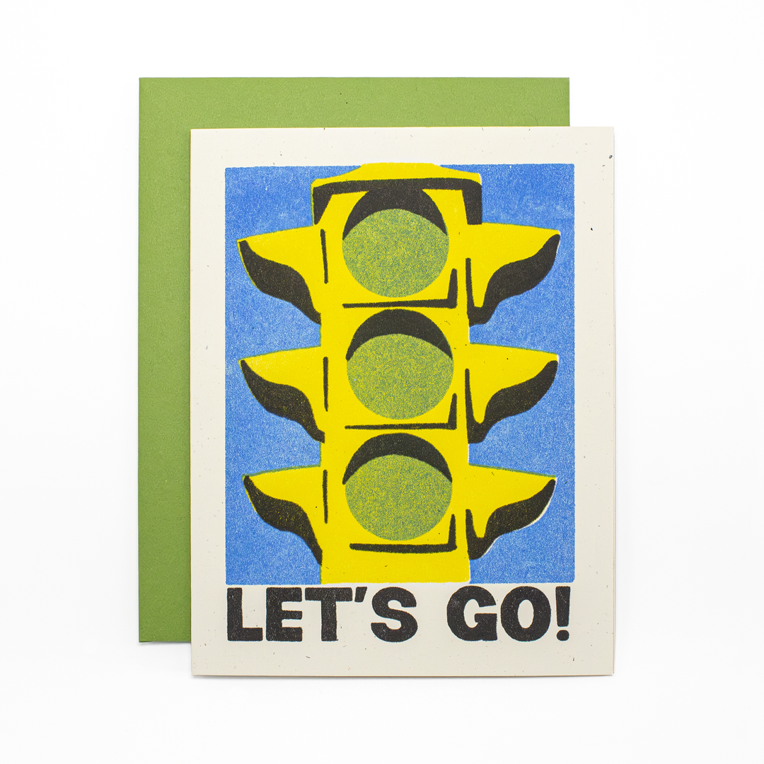 A greeting card with a bold graphic of a yellow traffic light on a blue background and the text 'LET'S GO!' in black, shown with a green envelope behind it.