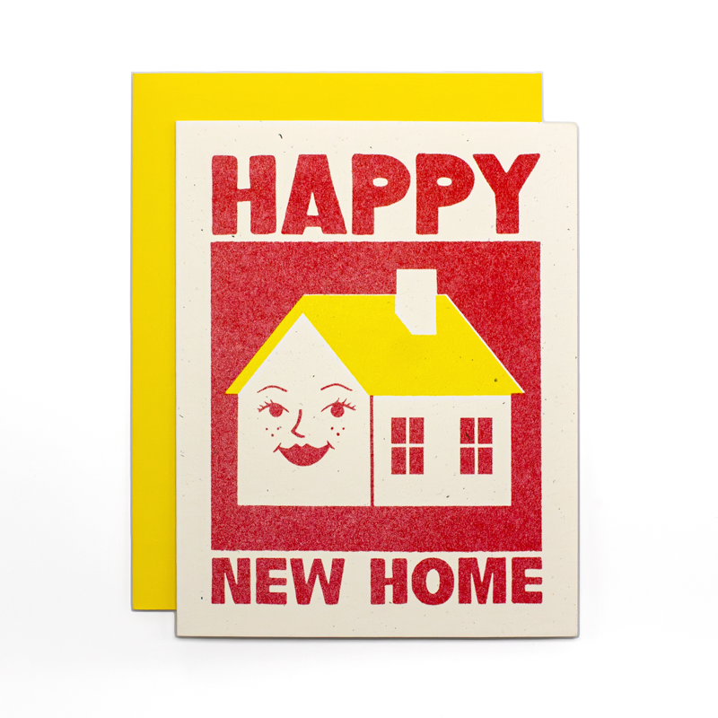 A greeting card with a bright yellow envelope behind it. The card has a vintage-style red and cream illustration of a house with a smiling face and the words “HAPPY NEW HOME” in bold red lettering.