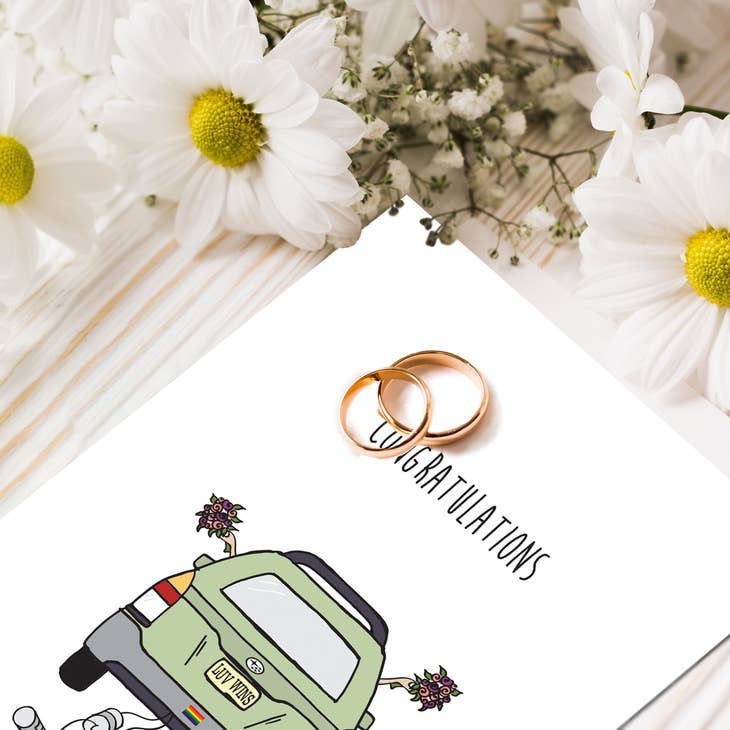 Greeting card with text Congratulations and drawing of car with a bouquet out each window and tin cans tied to the back. The license plate reads LUV WINS. There are two wedding rings lying on the card