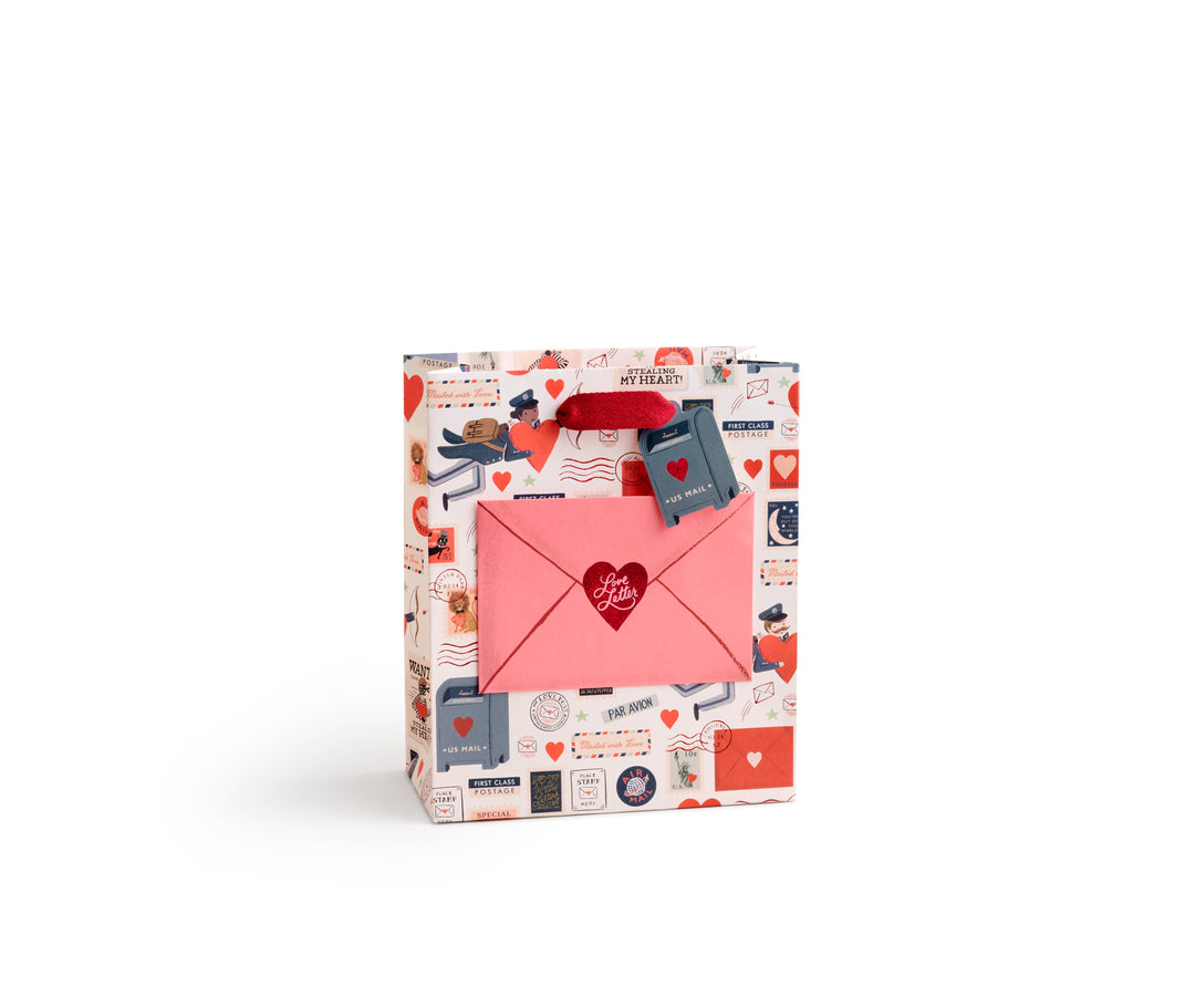 Gift bag with heart design and stationery-themed pattern with pink envelope in the center and a red grosgrain ribbon handle.