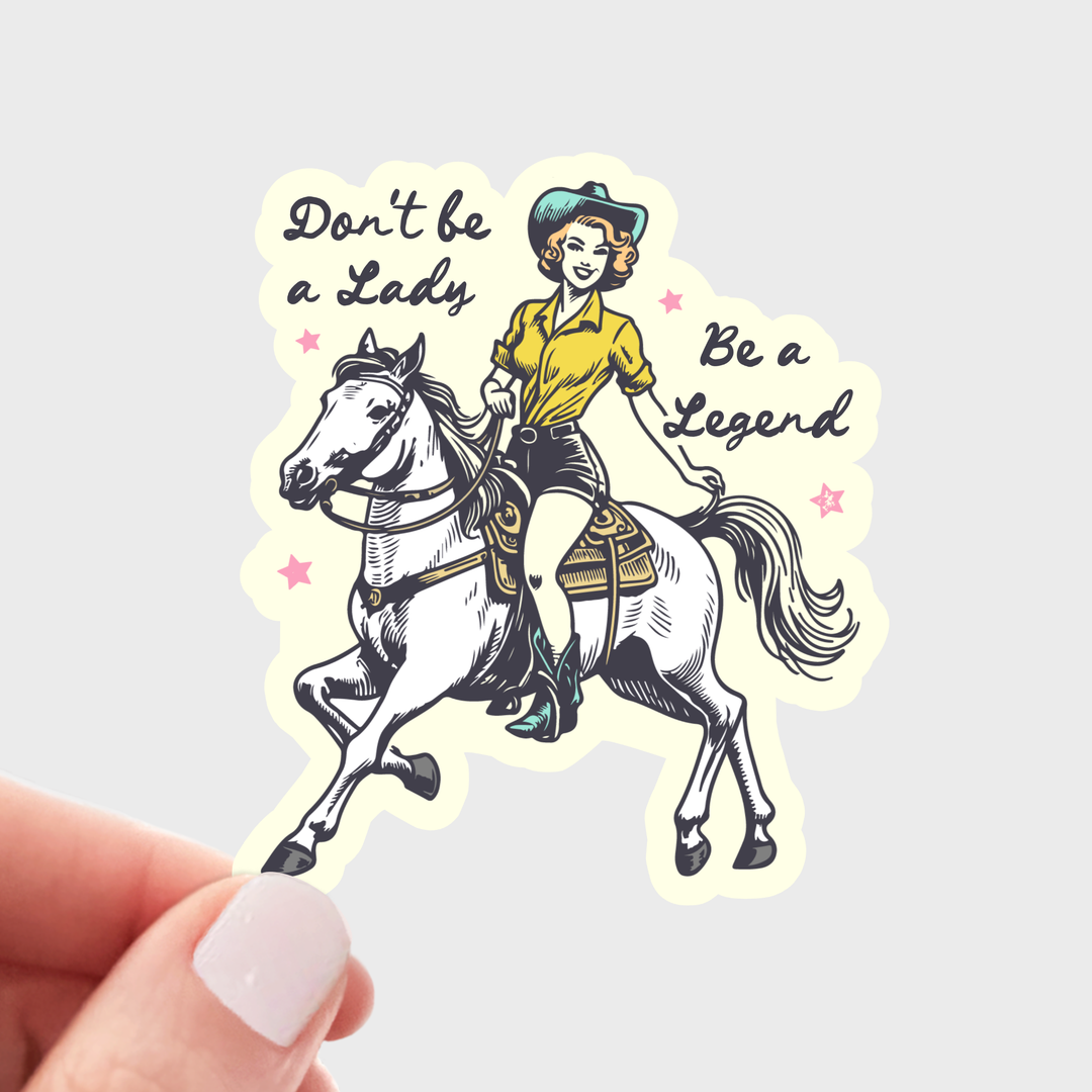 Hand holding a ticker with a woman wearing a blue cowboy hat, yellow shirt, shorts and cowboy boots, riding a white horse with text 'Don't be a lady, be a legend.'