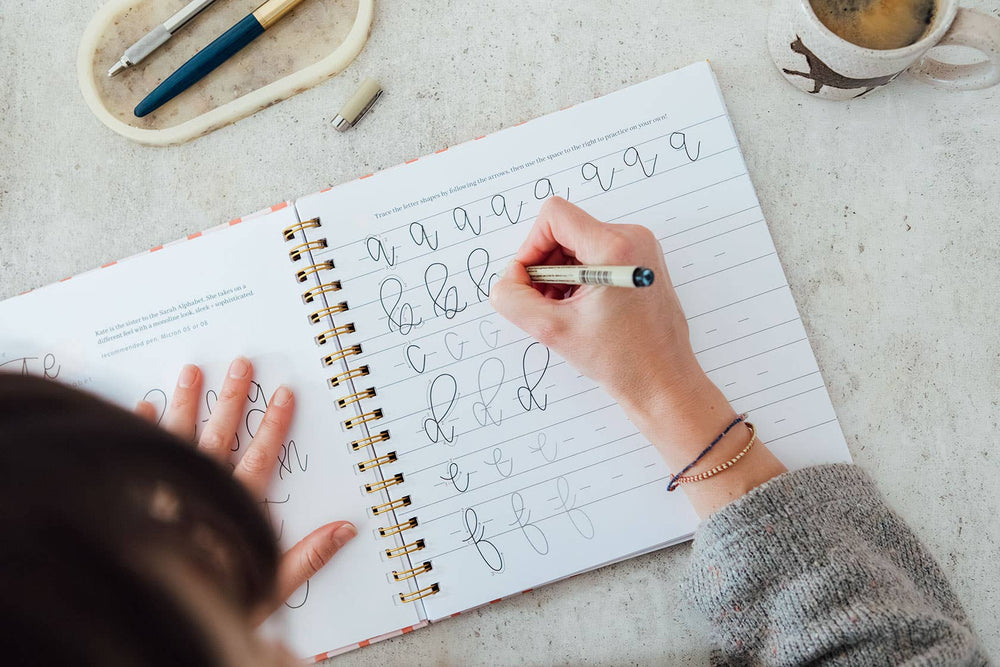 Person practicing calligraphy on a notebook with a pen and coffee mug in the background.