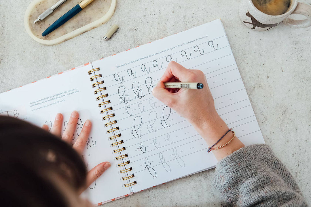 Person practicing calligraphy on a notebook with a pen and coffee mug in the background.
