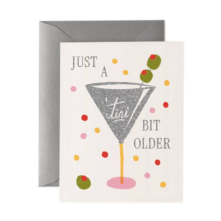 A glitter silver martini glass with two olives on a pick surrounded by festive colorful dots and a third olive. The text is placed around the glass with the word 'tini on the glass.