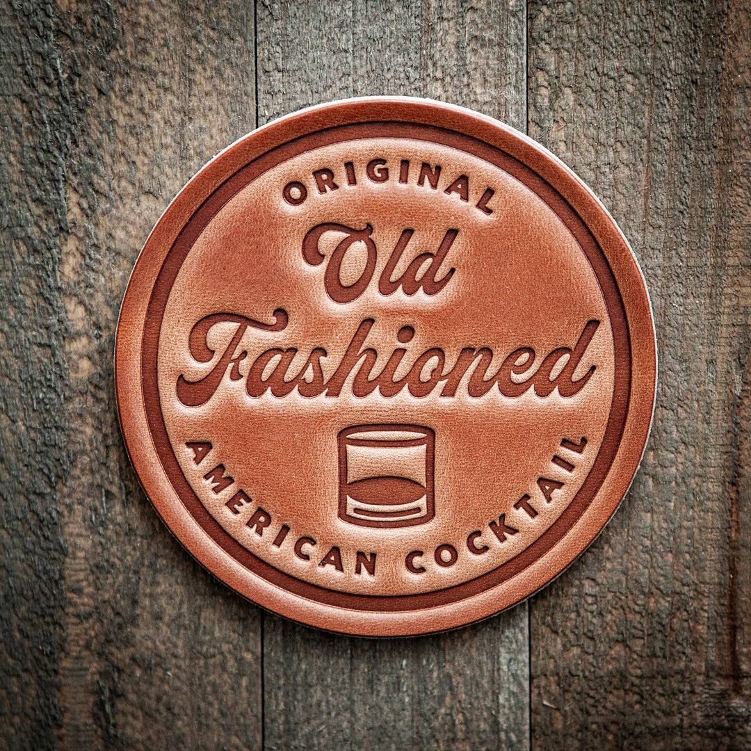 Round leather coaster  with 'Old Fashioned' text in script, a stylized illustration of an actual cocktail, and the words 'ORIGINAL AMERICAN COCKTAIL' around the circumference.