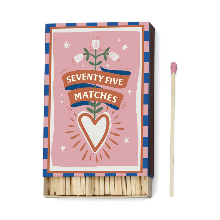 Partially opened match box whose whimsical illustration includes a white and brown heart, and a flower stem with three white blossoms. The border is alternating pink and blue rectangles.