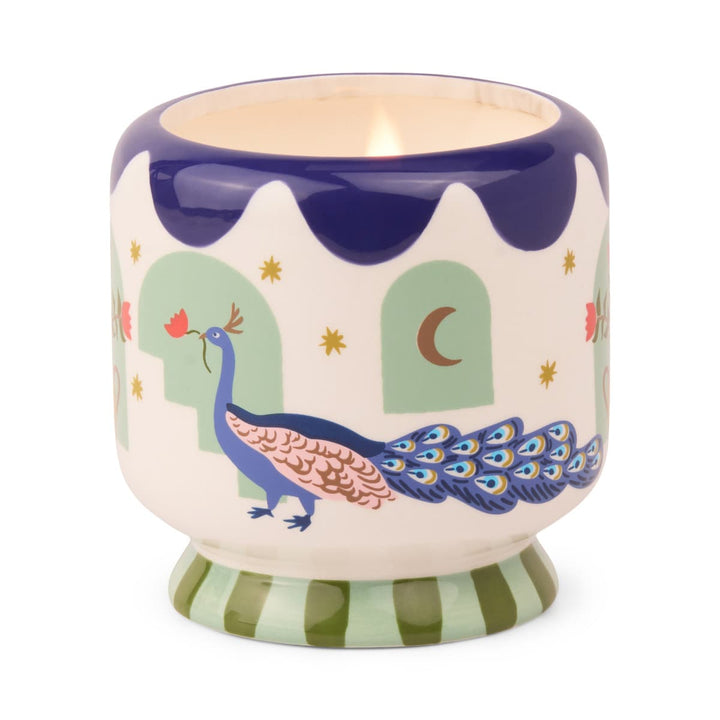 Footed ceramic vessel. The foot has alternating light and dark green stripes. The top is scalloped in navy blue. Around the outside is a predominately blue peacock holding a red flower and surrounded by a few gold stars and a quarter moon.