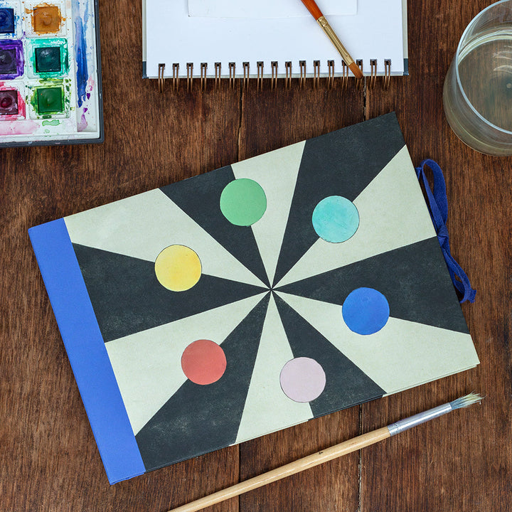 Sketchbook with colored circles on top of black and white geometric shapes; blue spine and closure ribbon.