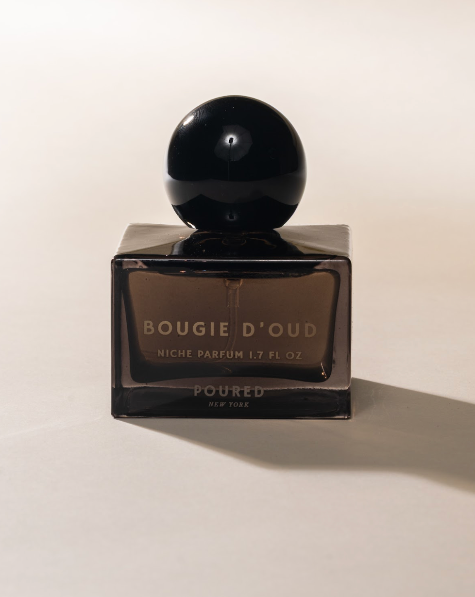 A dark amber glass perfume bottle with a glossy black spherical cap, labeled 'BOUGIE D'OUD' and 'POURED NEW YORK'.