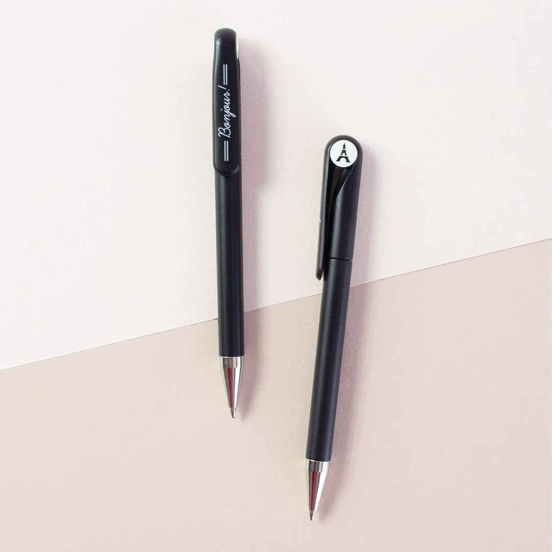 Two sides of a black retractable ballpoint pen with silver tips. One side shows the word 'Bonjour'' in white script, and the other side has a circular white cap insert with an image of the Eiffel  tower.