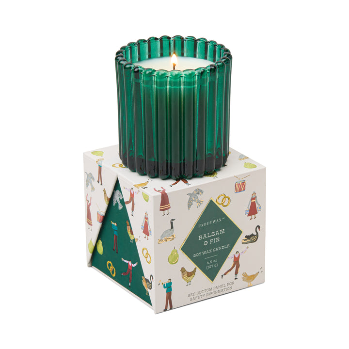 A green ribbed glass candle holder with a lit white candle, displayed on top of its illustrated white packaging box.