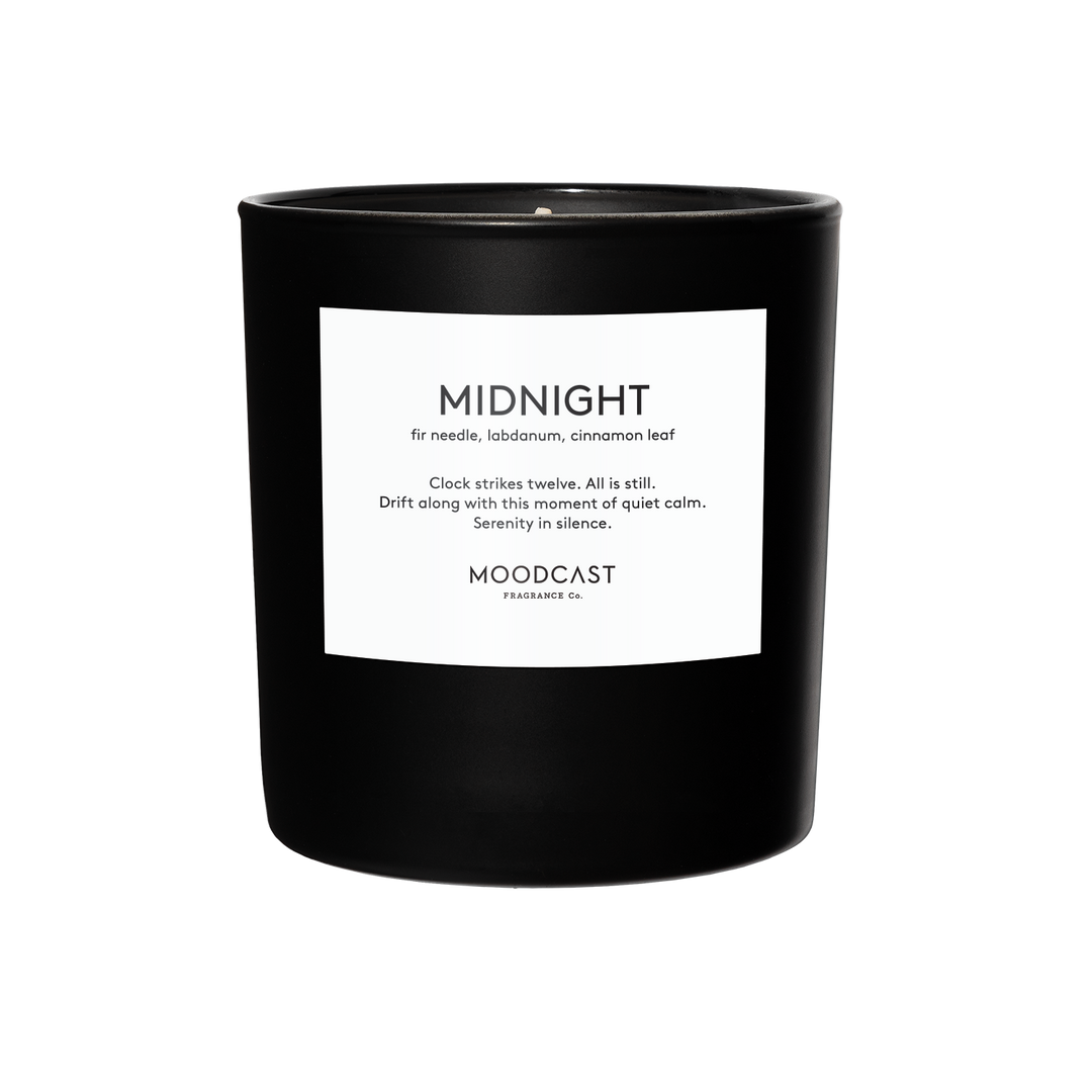 A black glass jar candle with a white label and blue text with the candle name 'MIDNIGHT', the scent profile, a description, and the brand name.