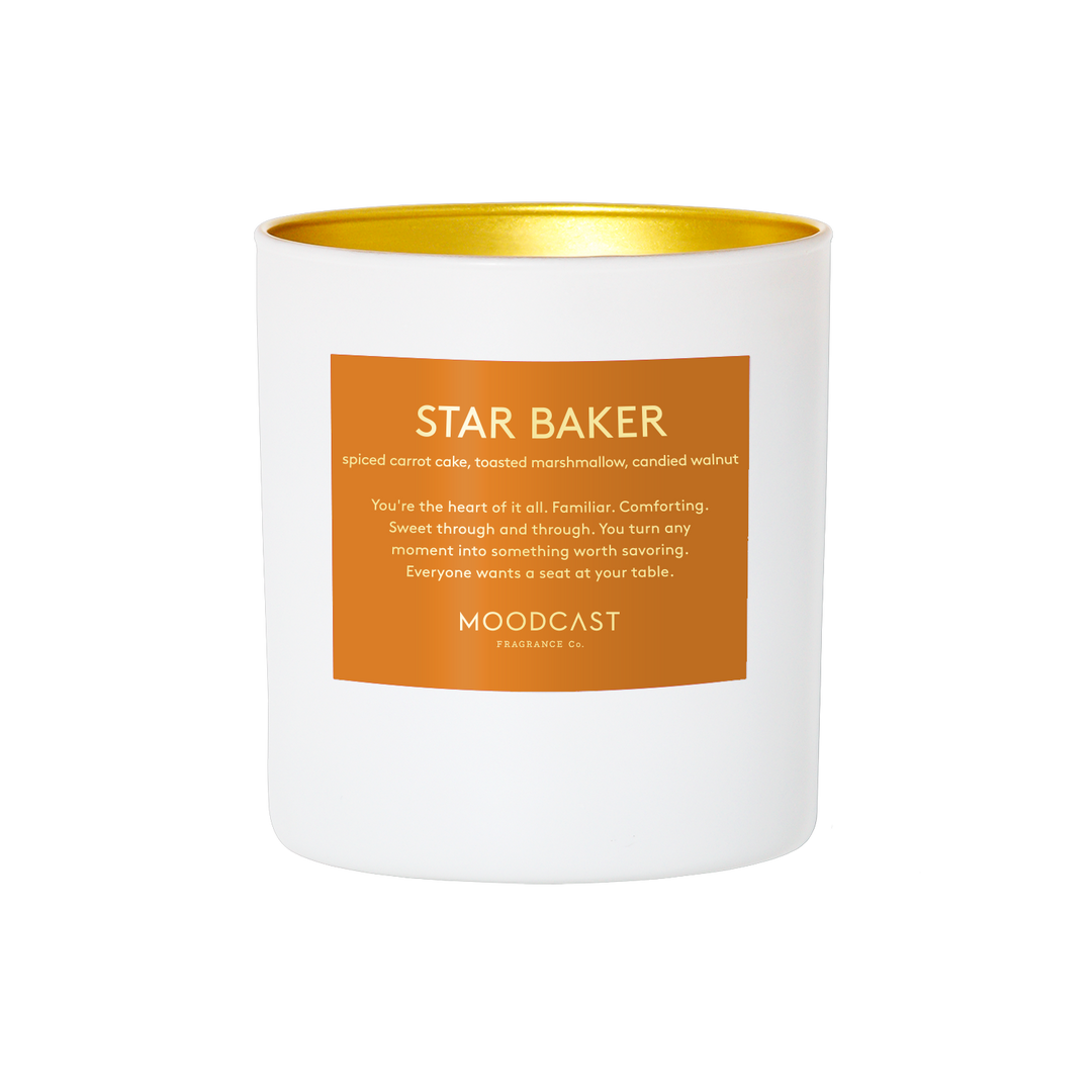 A white soy wax candle with a metallic gold interior and a square orange label on the front. The label features the scent name 'STAR BAKER' and the brand 'MOODCAST FRAGRANCE CO.
