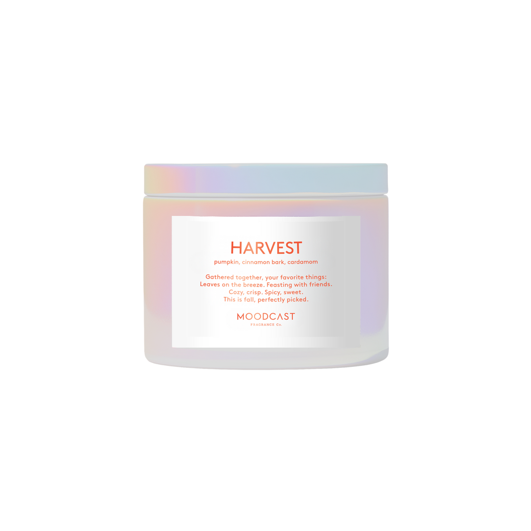 A jar of MOODCAST HARVEST candle, featuring a frosted white container with an iridescent pastel blue and pink lid, and a white label with orange text.