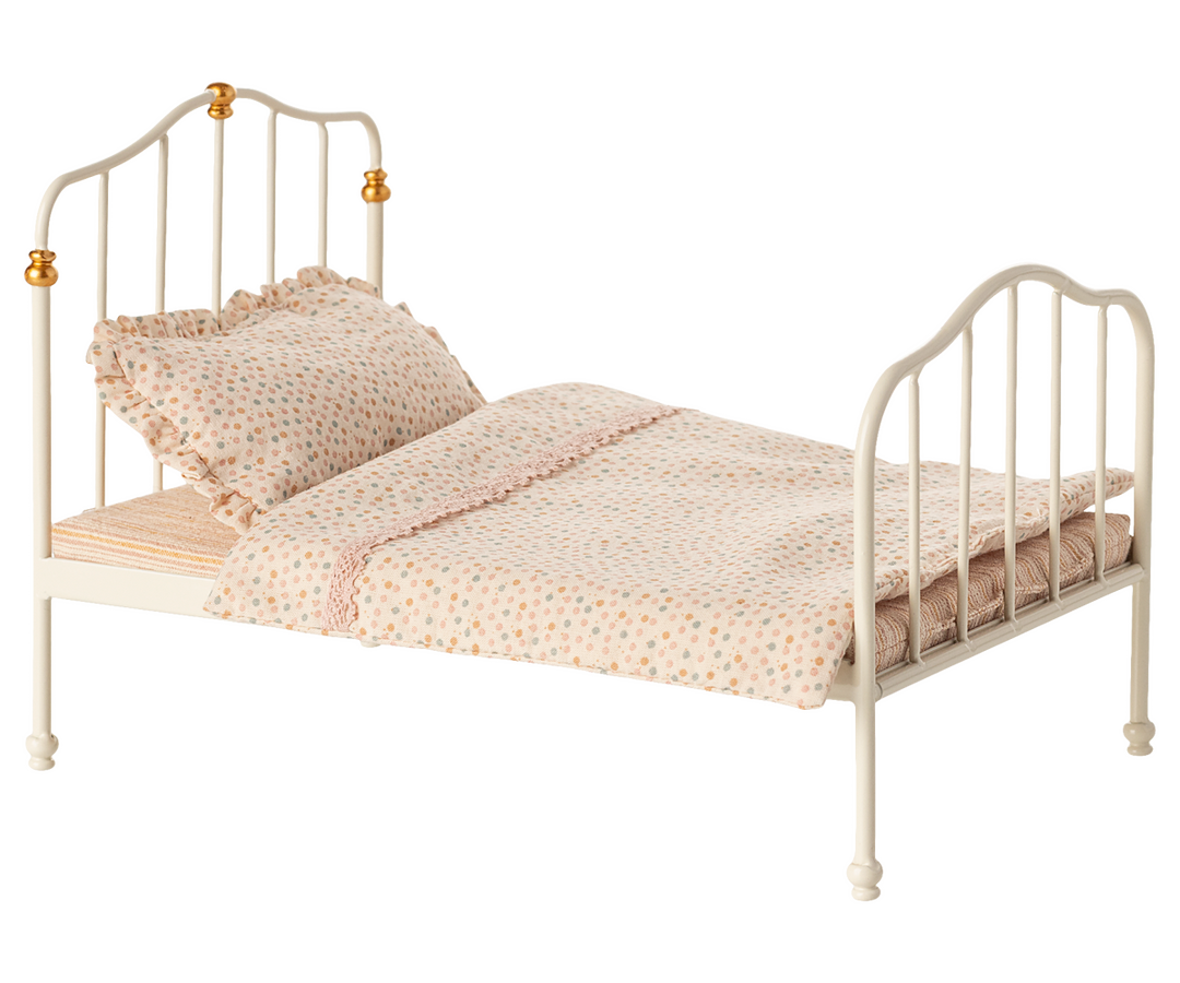 Bed with white metal frame with vertical bars at foot and head, and with a striped mattress, and dotted pillow and blanket. 