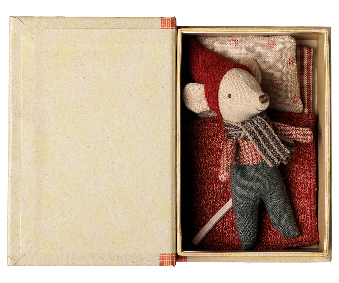 A small plush mouse toy dressed in a red hat and scarf with gray pants, displayed inside an open beige box with a red patterned pillow and red fabric mattress.
