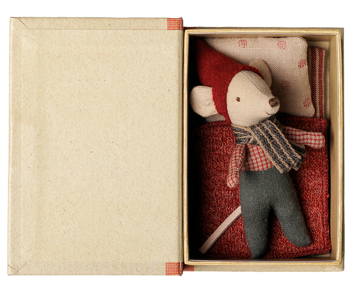 A small plush mouse toy dressed in a red hat and scarf with gray pants, displayed inside an open beige box with a red patterned pillow and red fabric mattress.