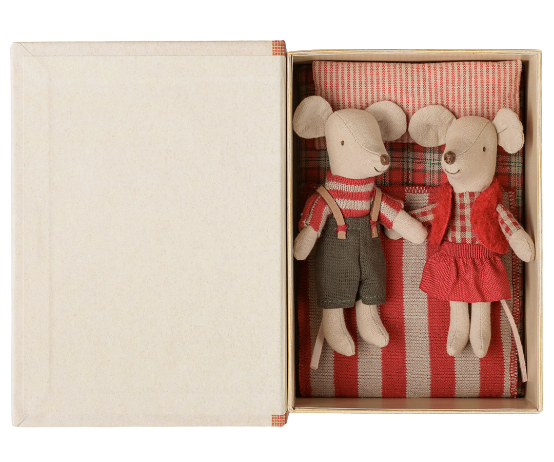 Mouse twins wearing their Christmas outfits inside the story book box lying on a red and off-white striped pillow, red-checked fabric mattress and red and off-white striped blanket.
