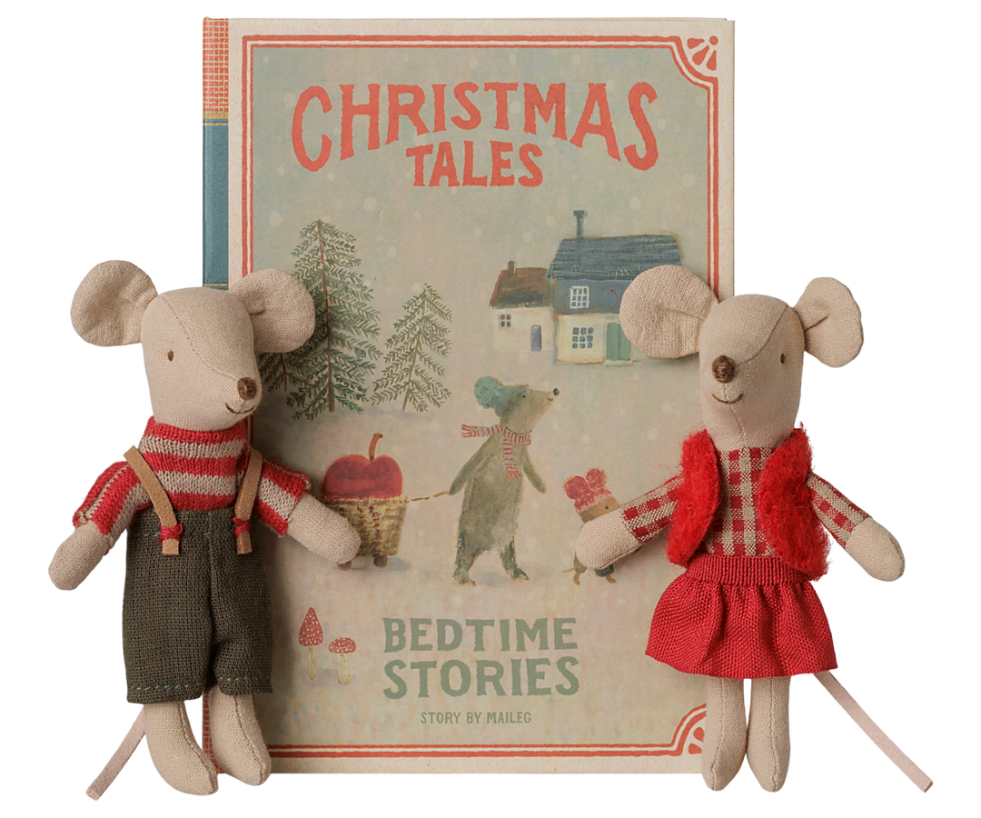 A vintage-style book titled 'Christmas Tales: Bedtime Stories' stands upright between two small mice dressed in holiday clothing. The book cover shows a snowy house scene with trees, and the mice are wearing holiday outfits: a red striped top and gray overalls for the boy and a red checked top, with red skirt and vest for the girl. 