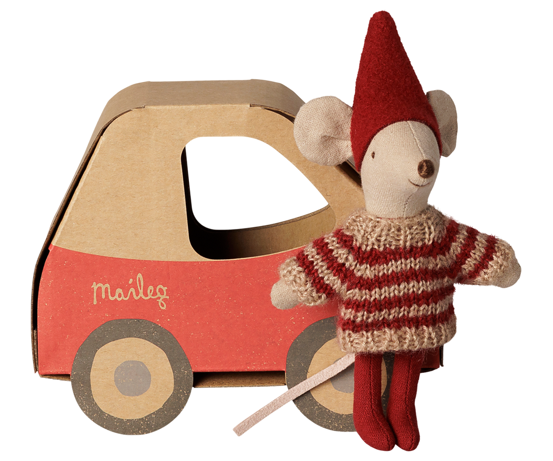 Little sister mouse wearing a red pixy hat, red and beige striped knit sweater, and red tights, standing next to a red and brown cardboard car with the brand name 'Maileg' printed on the side.