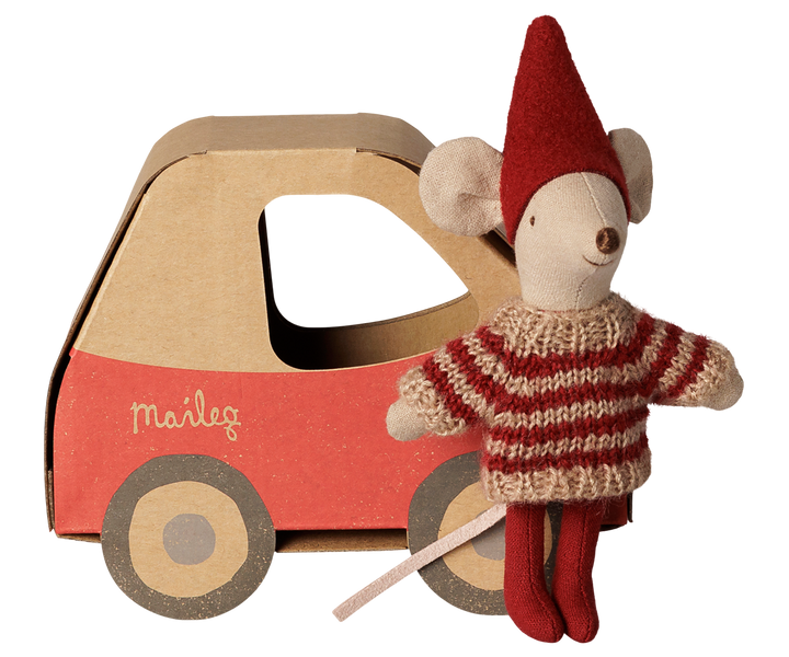 Little sister mouse wearing a red pixy hat, red and beige striped knit sweater, and red tights, standing next to a red and brown cardboard car with the brand name 'Maileg' printed on the side.