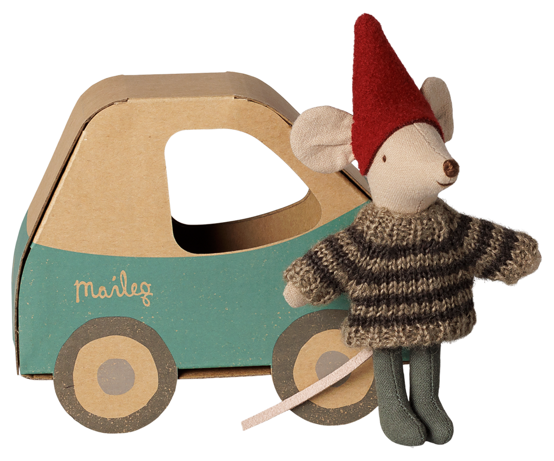 Little brother mouse wearing a red pixy hat, brown and beige striped knit sweater, and gray tights, standing next to a blue and brown cardboard car with the brand name 'Maileg' printed on the side.