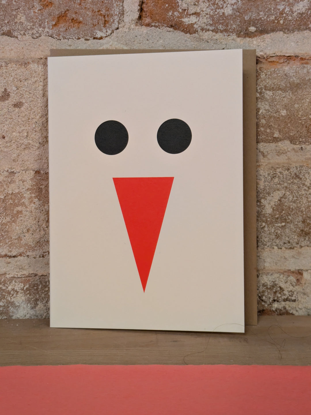 Abstract snowman with two large black circles for eyes and a red triangle for a nose on a white card.