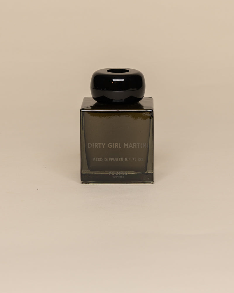A square dark brown glass reed diffuser bottle with a glossy black cap, labeled 'DIRTY GIRL MARTINI' and 'REED DIFFUSER 3.4 FL OZ'.