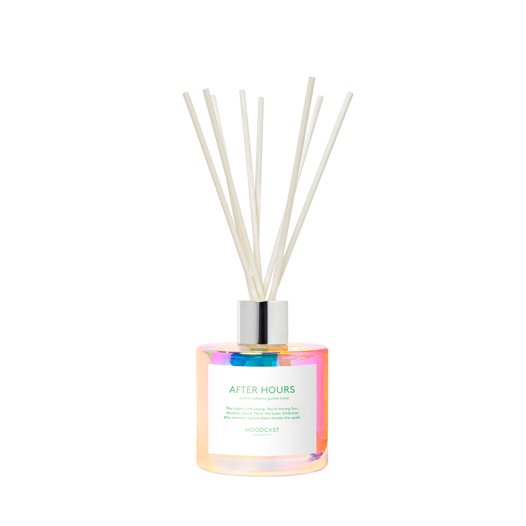 A reed diffuser bottle with an iridescent finish, a white label reading 'AFTER HOURS' and 'MOODCAST', and several natural reed sticks in a silver cap.