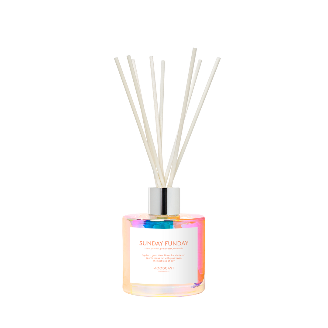 A reed diffuser bottle with an iridescent finish, a white label reading 'SUNDAY FUNDAY' and 'MOODCAST', and several off-white reed sticks in a silver cap.